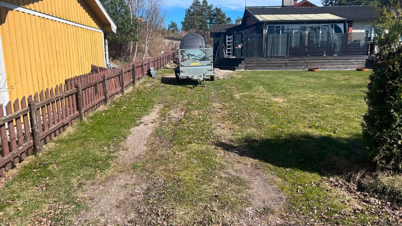 Trädgårdsyta före arbetet med leriga spår och till släpvagn, i bakgrunden gult förråd och mörkt bostadshus